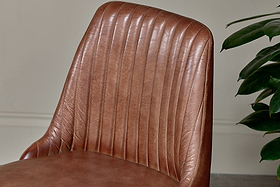 Close-up of nkuku’s Harsha Leather Dining Chair in chocolate brown, featuring smooth, ribbed buffalo leather upholstery and vertical stitching on the backrest. A neutral wall and green leafy plant appear in the background.