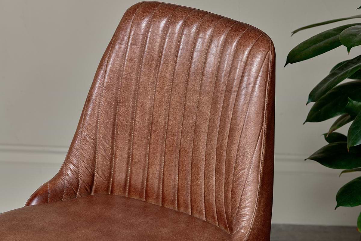 Close-up of nkuku’s Harsha Leather Dining Chair in chocolate brown, featuring smooth, ribbed buffalo leather upholstery and vertical stitching on the backrest. A neutral wall and green leafy plant appear in the background.
