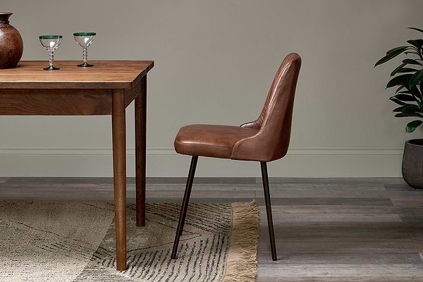 The Harsha Leather Dining Chair in chocolate brown by nkuku, with black metal legs, sits beside a wooden table holding two green-rimmed glasses. A patterned rug covers wood floors, and a potted plant rests against the neutral wall behind.
