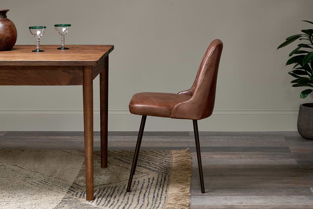 The Harsha Leather Dining Chair in chocolate brown by nkuku, with black metal legs, sits beside a wooden table holding two green-rimmed glasses. A patterned rug covers wood floors, and a potted plant rests against the neutral wall behind.
