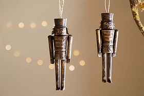 The Imay Nutcracker Baubles - Antique Brass (Set of 2) by nkuku feature simple rectangular soldiers with tall hats, hanging from strings—ideal festive decor for your Christmas tree against a softly lit background.