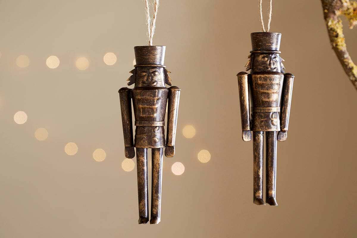 The Imay Nutcracker Baubles - Antique Brass (Set of 2) by nkuku feature simple rectangular soldiers with tall hats, hanging from strings—ideal festive decor for your Christmas tree against a softly lit background.