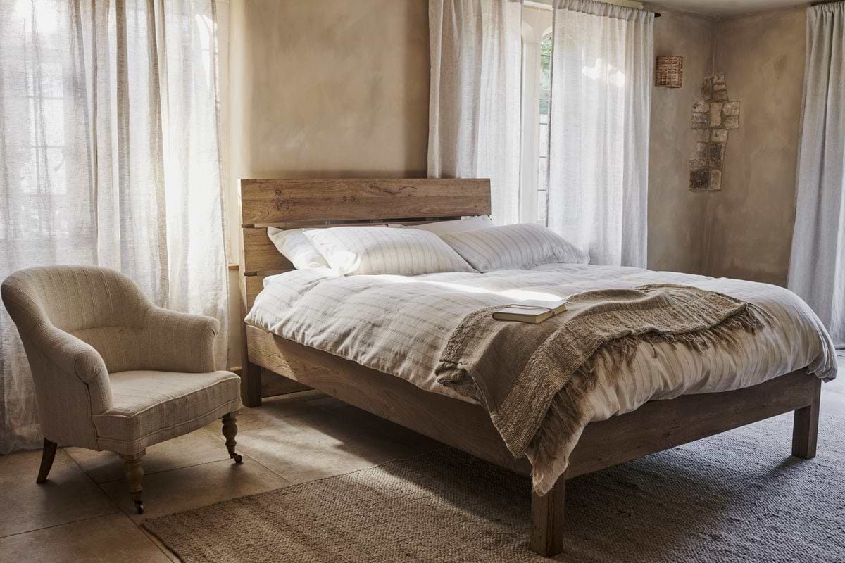 A cozy bedroom featuring the nkuku Iya Reclaimed Wood Bed in natural, white and beige bedding, an open book, an upholstered armchair, large windows with sheer curtains, and soft natural light.