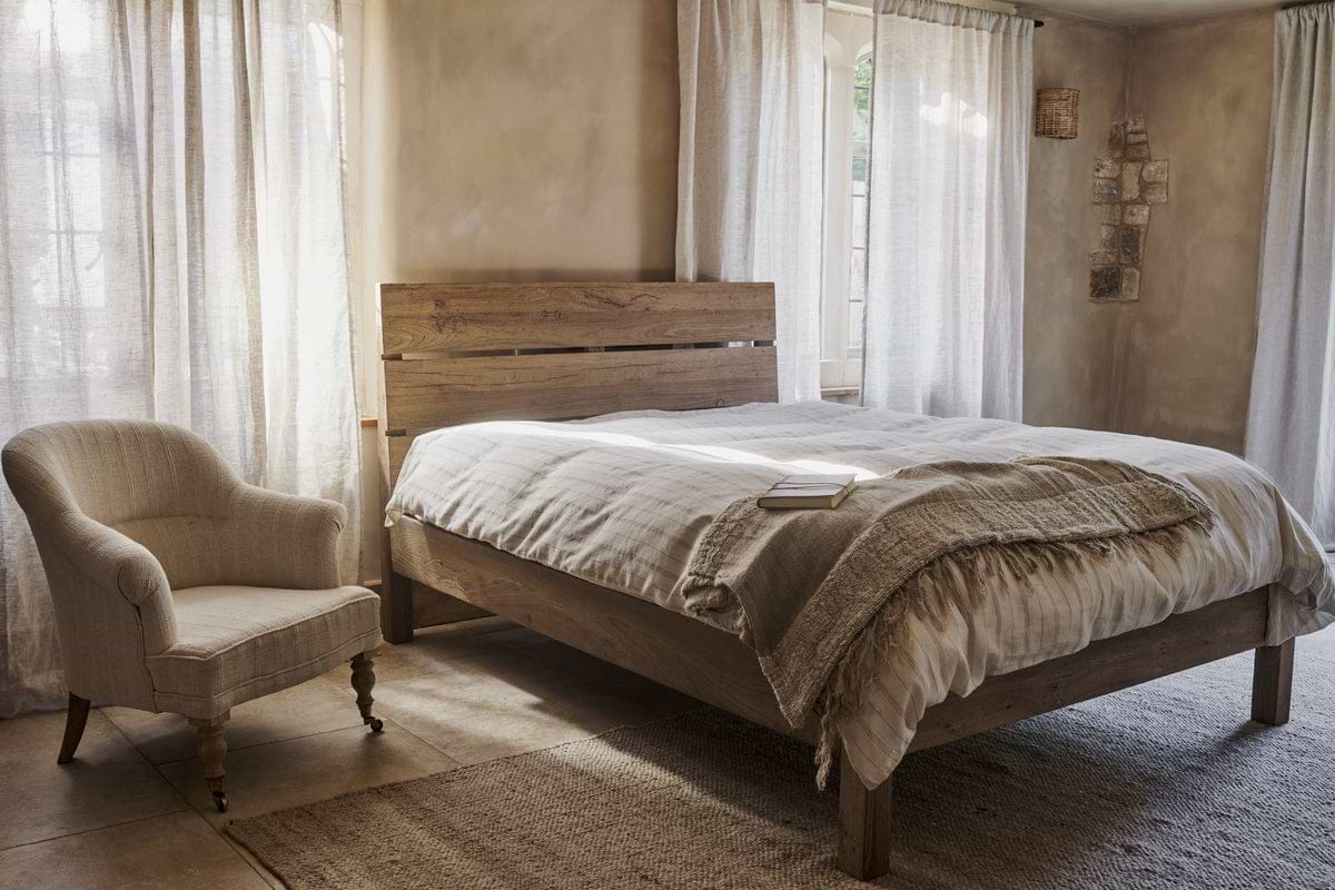 A cozy bedroom features the nkuku Iya Reclaimed Wood Bed - Natural, neutral bedding, an upholstered armchair, soft light from large windows with sheer curtains, and a textured rug. A book rests on the bed.