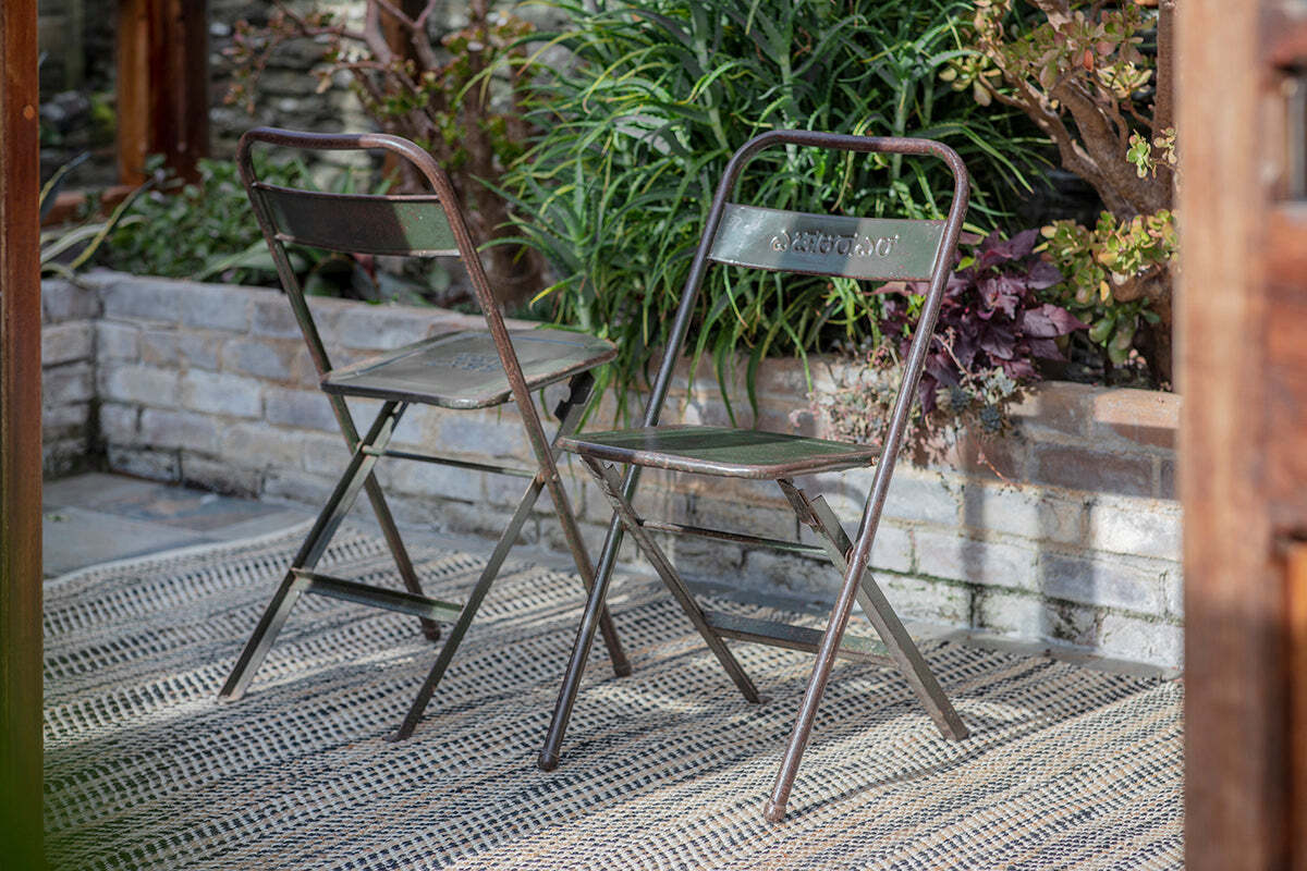 Two Ishan Reclaimed Folding Chairs by nkuku sit on a woven outdoor rug in a garden patio, facing slightly apart amid green leafy plants and a low brick wall. Sunlight casts gentle shadows, enhancing the rustic charm of the scene.