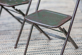 A close-up of two nkuku Ishan Reclaimed Folding Chairs, showing their aged metal frames and faded green seats on a zigzag rug. The worn texture and rustic look highlight the chair’s nostalgic charm.