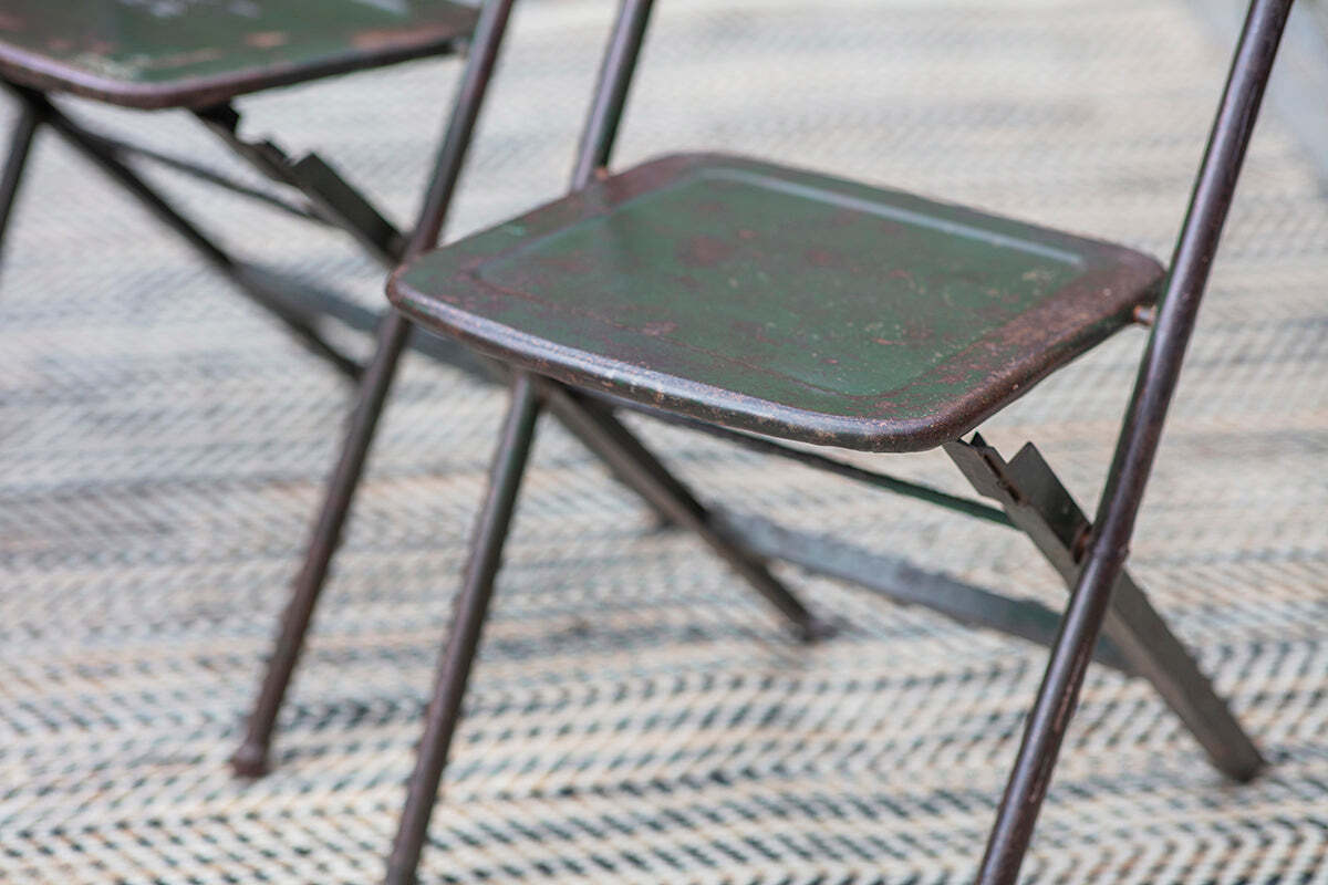 A close-up of two nkuku Ishan Reclaimed Folding Chairs, showing their aged metal frames and faded green seats on a zigzag rug. The worn texture and rustic look highlight the chair’s nostalgic charm.
