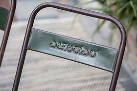 A close-up of the green-painted backrest of the Ishan Reclaimed Folding Chair by nkuku, embossed with Kannada script reading ಎಜ್ಜರಬುವುದು, highlights its rustic wear against a blurred plant in the background.