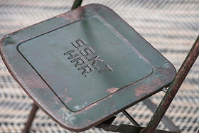 A close-up of the Ishan Reclaimed Folding Chair by nkuku shows its weathered green finish, rust details, and embossed S.S.K.T H.R.R. text on the seat. It sits on a patterned woven surface, adding rustic charm to the setting.