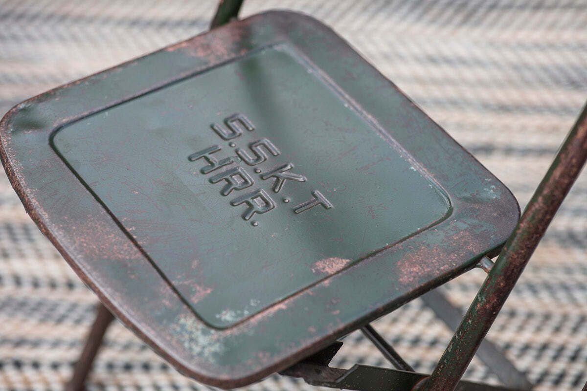 A close-up of the Ishan Reclaimed Folding Chair by nkuku shows its weathered green finish, rust details, and embossed S.S.K.T H.R.R. text on the seat. It sits on a patterned woven surface, adding rustic charm to the setting.