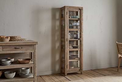 The nkuku Ibo Reclaimed Wood & Glass Cabinet - Natural - Tall, with antique brass handles, showcases glassware and bowls in a cozy room featuring a wooden floor, sideboard with dishes, and a wicker chair.