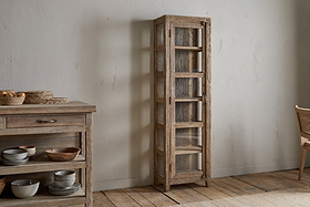 The Ibo Reclaimed Wood & Glass Cabinet - Natural - Tall by nkuku stands against a light wall on a wooden floor beside a matching sideboard adorned with ceramic dishes and woven baskets.