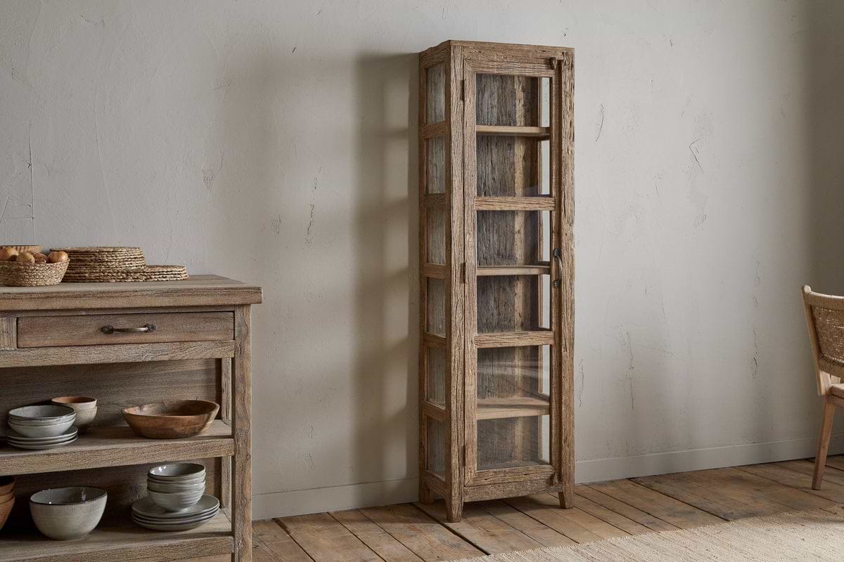 The Ibo Reclaimed Wood & Glass Cabinet - Natural - Tall by nkuku stands against a light wall on a wooden floor beside a matching sideboard adorned with ceramic dishes and woven baskets.