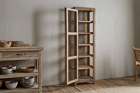 The nkuku Ibo Reclaimed Wood & Glass Cabinet - Natural - Tall stands open beside a rustic cabinet filled with bowls and baskets in a minimalist room featuring wooden floors and neutral walls.