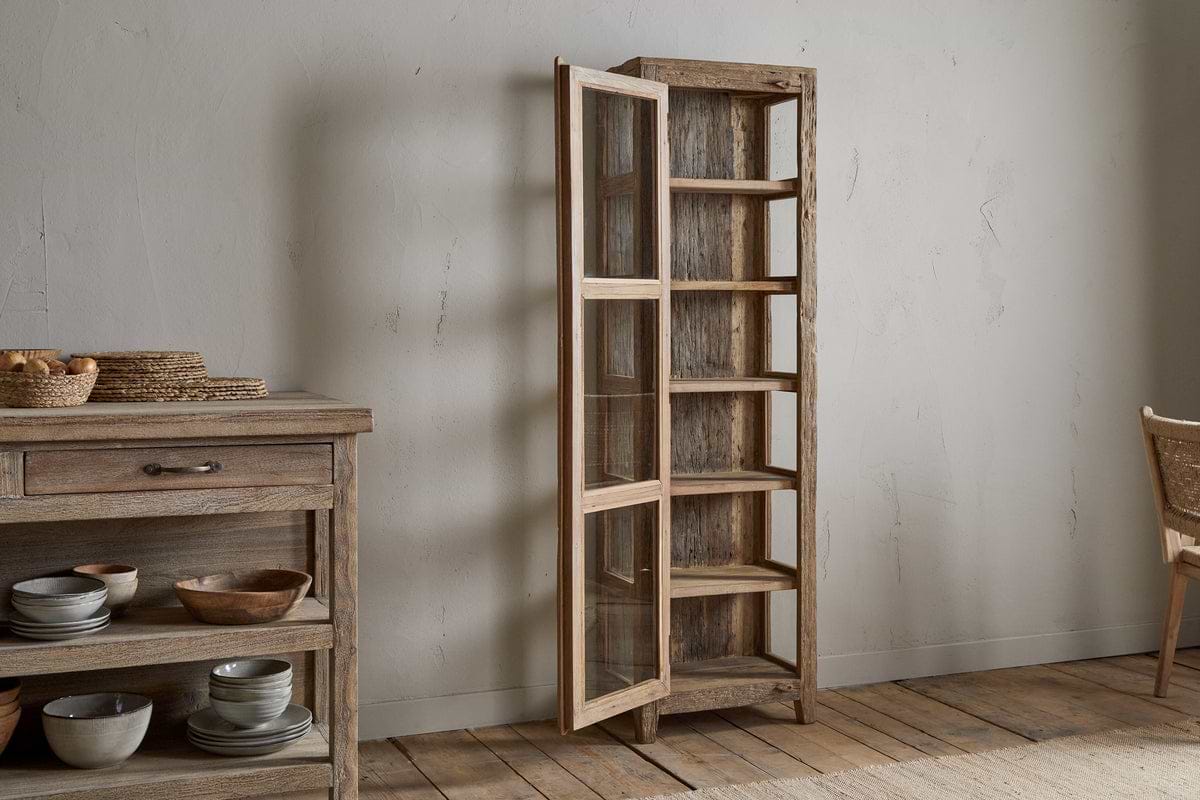 The nkuku Ibo Reclaimed Wood & Glass Cabinet - Natural - Tall stands open beside a rustic cabinet filled with bowls and baskets in a minimalist room featuring wooden floors and neutral walls.