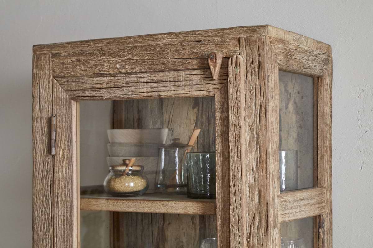 The nkuku Ibo Reclaimed Wood & Glass Cabinet - Natural - Tall features weathered wood, glass panels, antique brass handles, and displays glass jars, a bowl, and kitchen items on its upper shelves for a charming vintage look.