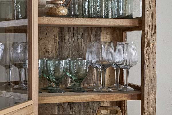 The nkuku Ibo Reclaimed Wood & Glass Cabinet (Natural, Tall) showcases green and clear textured goblets, a cork-lidded jar, and a wooden cutting board on its rustic wooden shelves behind antique brass handles.