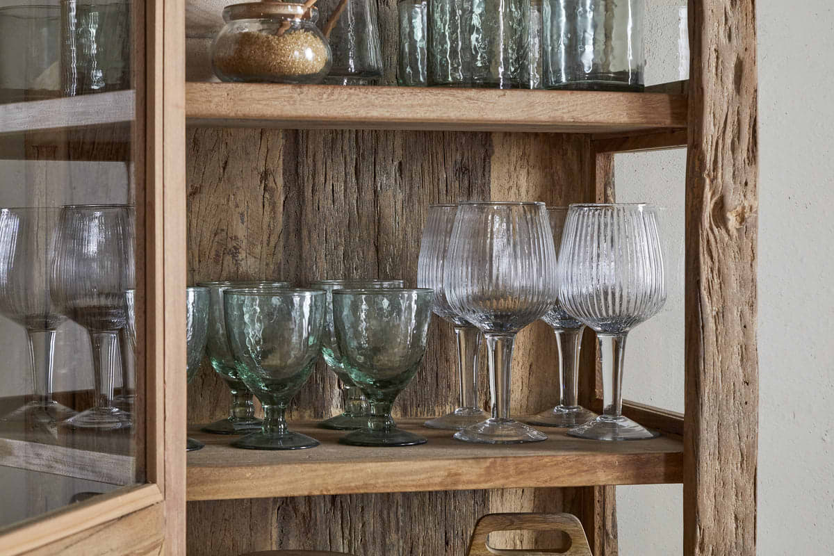The nkuku Ibo Reclaimed Wood & Glass Cabinet (Natural, Tall) showcases green and clear textured goblets, a cork-lidded jar, and a wooden cutting board on its rustic wooden shelves behind antique brass handles.