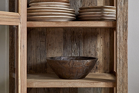 The nkuku Ibo Reclaimed Wood & Glass Cabinet - Natural - Tall features antique brass handles, stacked wooden plates on the top shelf, and a carved wooden bowl on the middle shelf against a weathered wood back panel.