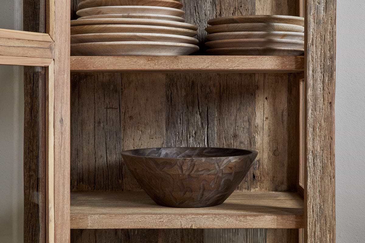 The nkuku Ibo Reclaimed Wood & Glass Cabinet - Natural - Tall features antique brass handles, stacked wooden plates on the top shelf, and a carved wooden bowl on the middle shelf against a weathered wood back panel.
