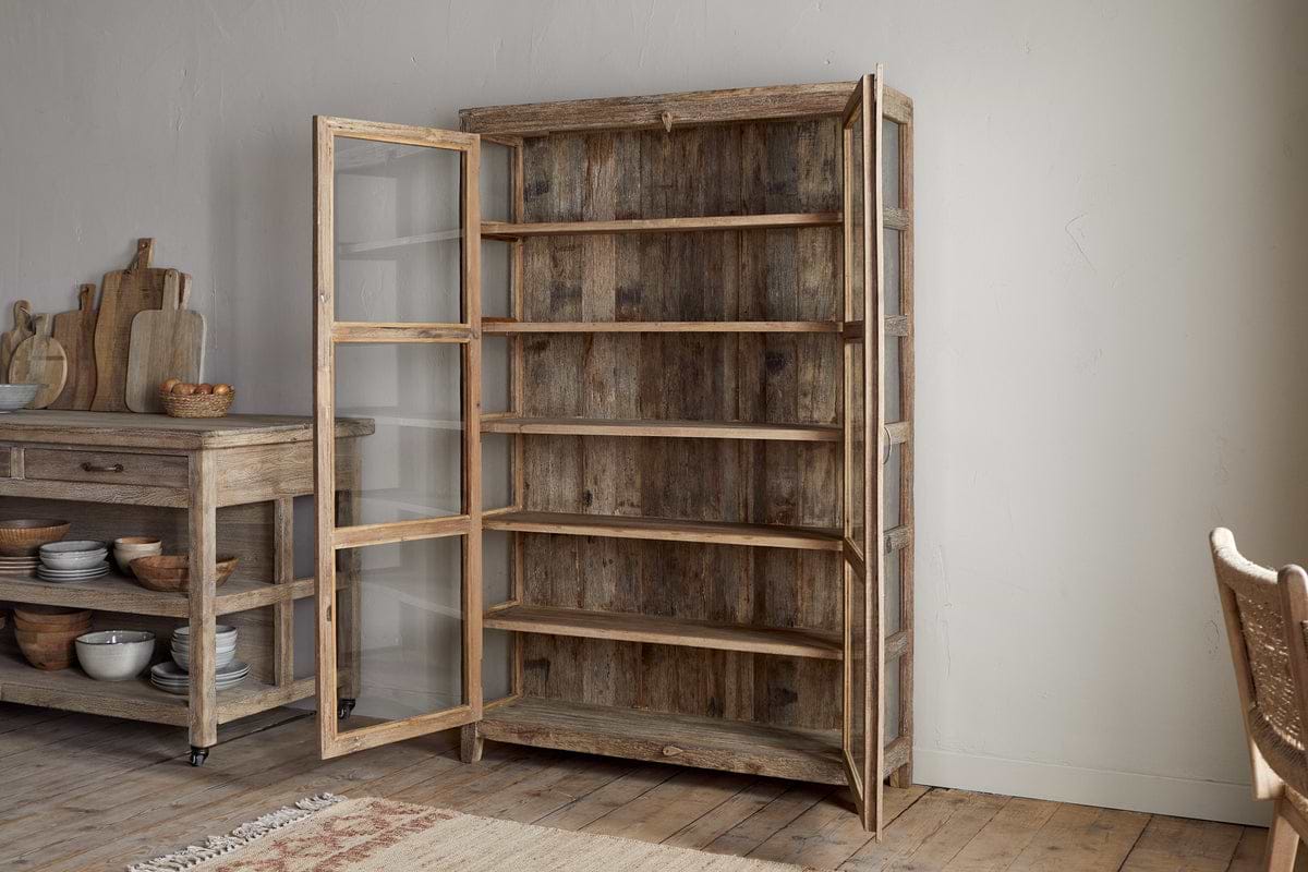 The nkuku Ibo Reclaimed Wood & Glass Cabinet - Natural - Wide features antique brass handles and glass panels, stands open with five shelves, and adds rustic charm to a cozy kitchen with wooden floors and light walls.