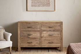 The Iya Reclaimed Wood Chest of Drawers - Natural - Large by nkuku, featuring six drawers with metal handles, stands against a light wall beneath textured artwork, flanked by a beige armchair and a pink-covered bed.