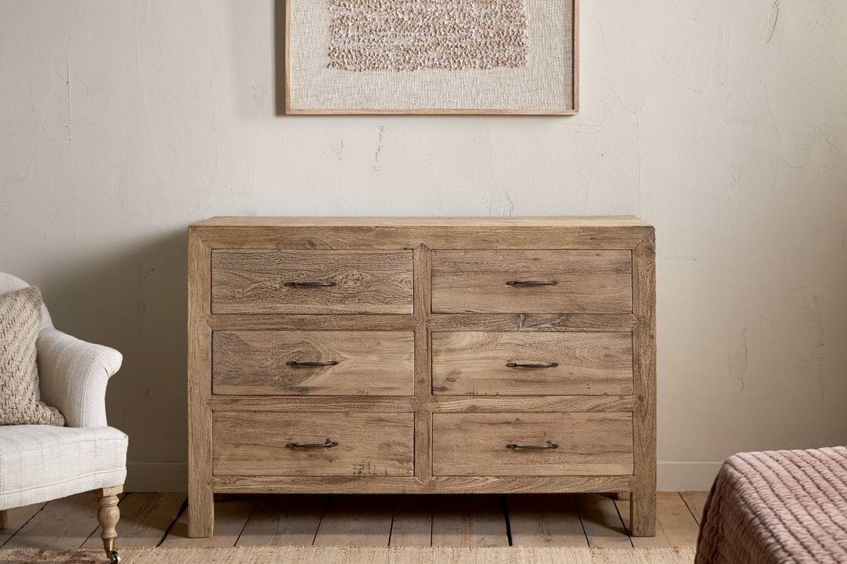 The Iya Reclaimed Wood Chest of Drawers - Natural - Large by nkuku, featuring six drawers with metal handles, stands against a light wall beneath textured artwork, flanked by a beige armchair and a pink-covered bed.