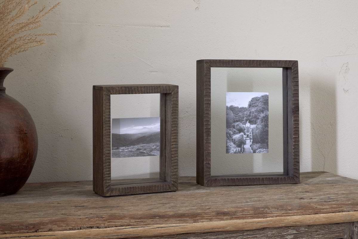 Two nkuku Indu Standing Mango Wood Frames in Dark Ash, each displaying black and white landscape photos, rest on a rustic wooden surface against a light wall. On the left, a brown vase with dried grass is partially visible.