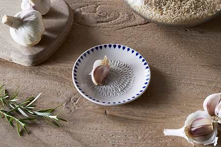A wooden table features nkukus Indigo Drop Garlic Grater in Cream & Indigo with a clove of garlic, surrounded by garlic bulbs, rosemary sprig, a wooden board, and a glass jar of brown rice.