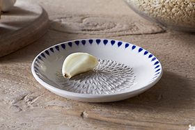 The nkuku Indigo Drop Garlic Grater in cream and indigo, featuring a textured center and hand-painted design, sits on a small white plate—perfect for grating garlic and adding style to your kitchen.