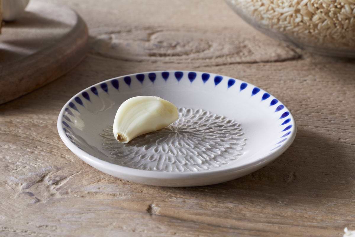 The nkuku Indigo Drop Garlic Grater in cream and indigo, featuring a textured center and hand-painted design, sits on a small white plate—perfect for grating garlic and adding style to your kitchen.