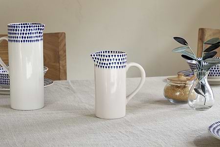 A small nkuku Indigo Drop Jug in cream and indigo sits on a linen tablecloth, surrounded by matching contemporary serveware, a sugar jar, and a glass vase filled with green leaves.
