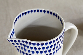 The nkuku Indigo Drop Jug - Cream & Indigo - Small features blue teardrop patterns along the rim. This white ceramic jug with a spout is ideal for contemporary serveware, displayed here on a neutral surface.
