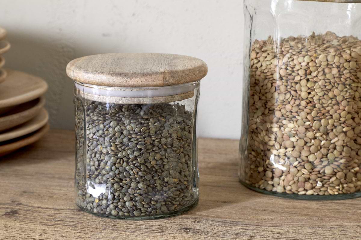 Two Igoko Storage Jars - Clear by nkuku, each with mango wood lids and filled with green and brown lentils, rest on a wooden surface beside stacked plates—an elegant way to organize your pantry.