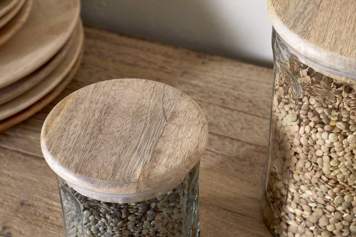 Two nkuku Igoko Storage Jars - Clear, with mango wood lids and filled with lentils, sit on a wooden surface beside stacked wooden plates, creating an elegant kitchen organizer.