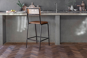 The Iswa Leather & Cane Counter Chair by nkuku, featuring an aged tan leather seat and woven cane back, stands on a herringbone wooden floor in a modern kitchen.