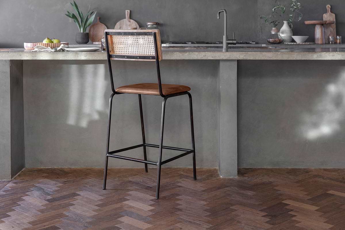 The Iswa Leather & Cane Counter Chair by nkuku, featuring an aged tan leather seat and woven cane back, stands on a herringbone wooden floor in a modern kitchen.