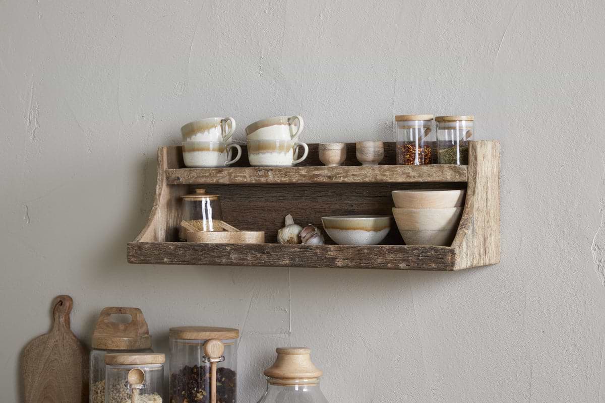 The nkuku Ibo Reclaimed Wood Storage Shelf in Natural hangs on a light wall, displaying ceramic cups, bowls, and spice jars above, with weathered wooden shelves below for extra kitchen storage of glass jars and a cutting board.