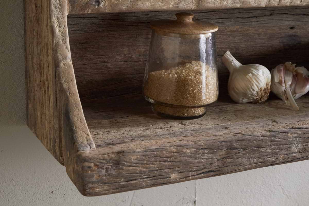 The Ibo Reclaimed Wood Storage Shelf by nkuku holds a glass jar with a wooden lid filled with brown sugar, plus garlic bulbs and cloves—ideal for stylish kitchen storage in a rustic setting.