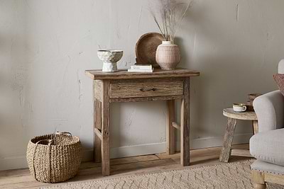 The nkuku Ibo Reclaimed Wood 1 Drawer Console Table in Natural brings rustic charm to your entryway, styled with a ceramic bowl, book, wooden tray, and vase with dried grasses. A woven basket sits nearby next to a cozy gray armchair.