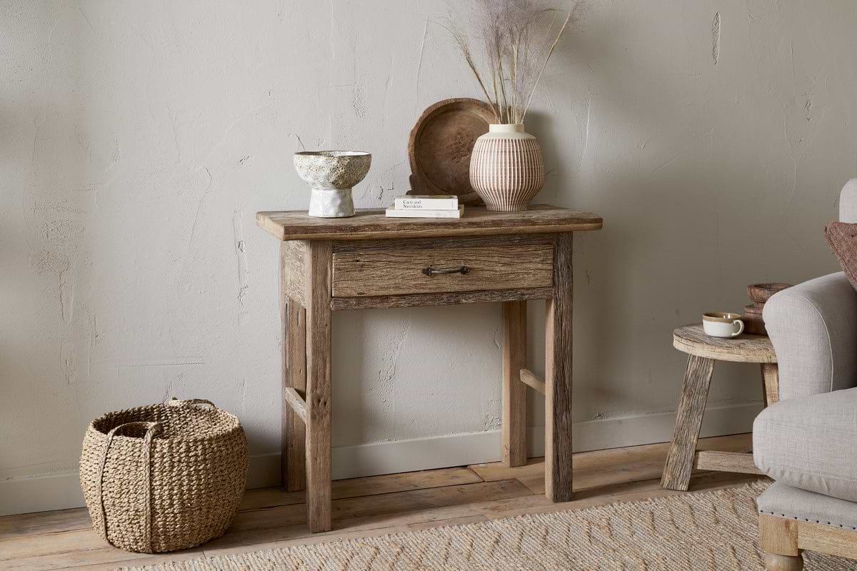 The nkuku Ibo Reclaimed Wood 1 Drawer Console Table in Natural brings rustic charm to your entryway, styled with a ceramic bowl, book, wooden tray, and vase with dried grasses. A woven basket sits nearby next to a cozy gray armchair.