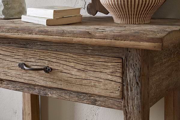 A close-up of the nkuku Ibo Reclaimed Wood 1 Drawer Console Table in Natural, showcasing a textured drawer with metal handle and weathered finish. Stacked books and a striped ceramic vase sit on top, set against a light entryway wall.