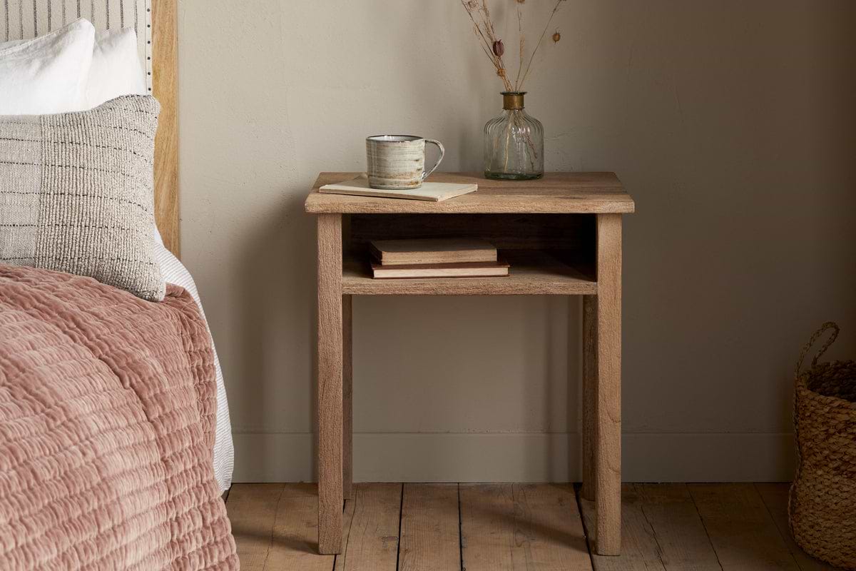 The Iya Reclaimed Wood Bedside Table by nkuku in natural holds a mug, book, and small glass vase with dried flowers. Two closed books sit on the shelf below. Nearby: a bed with a pink quilt and a woven basket create a cozy bedroom scene.