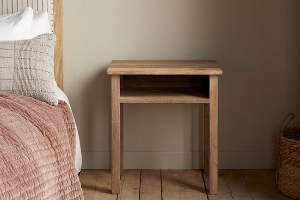 The Iya Reclaimed Wood Bedside Table by nkuku stands beside a bed with textured bedding and pillows. A woven basket rests on the wooden floor, creating a cozy bedroom scene against a plain beige wall.