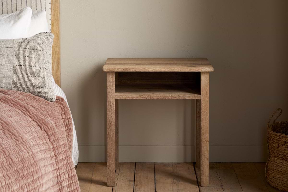 The Iya Reclaimed Wood Bedside Table by nkuku stands beside a bed with textured bedding and pillows. A woven basket rests on the wooden floor, creating a cozy bedroom scene against a plain beige wall.