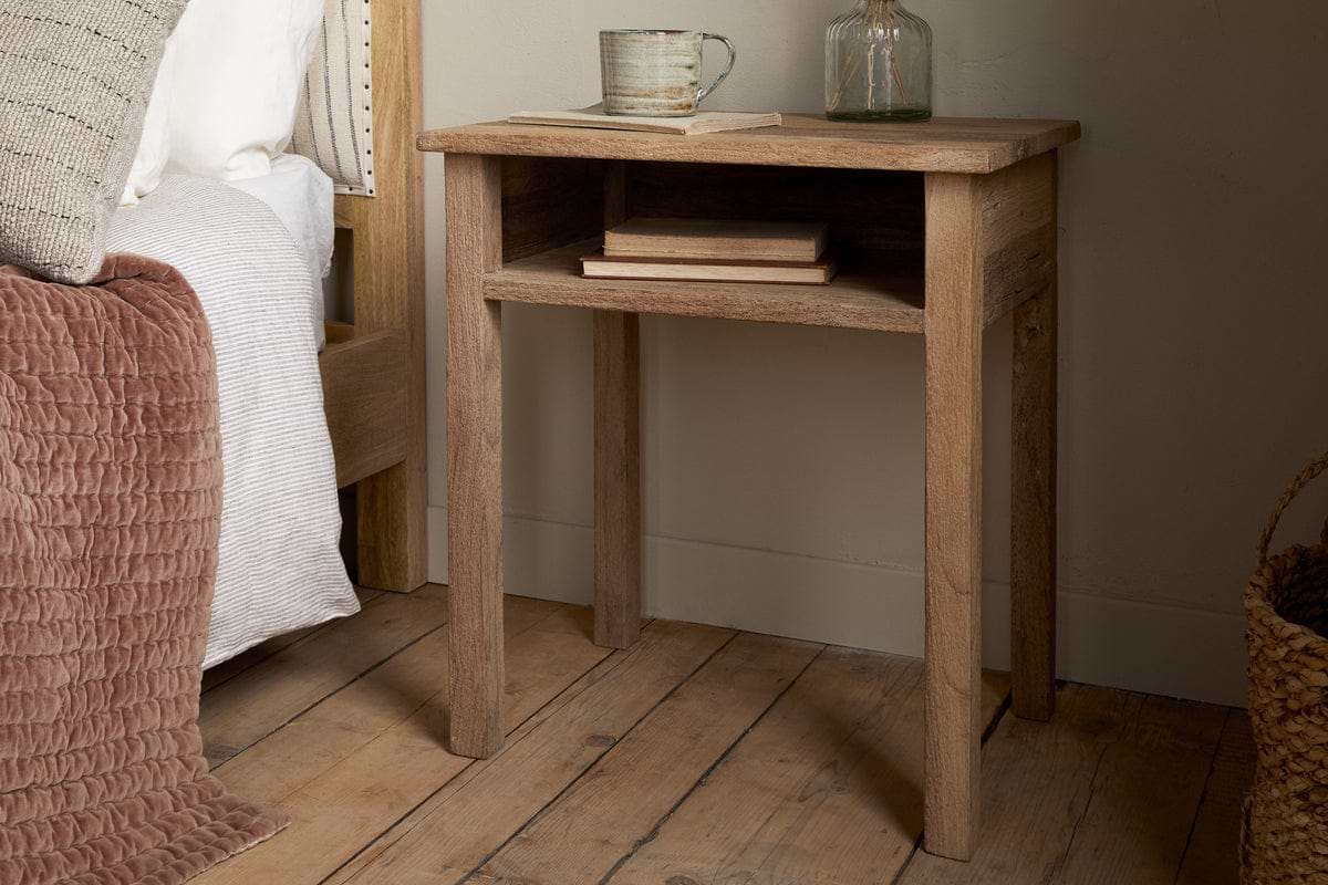 The Iya Reclaimed Wood Bedside Table by nkuku features an open shelf holding two books, a ceramic mug, and a glass bottle. It stands on a wooden floor beside a bed with white bedding and a textured dusty rose blanket.