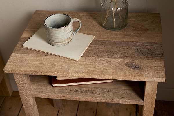 The Iya Reclaimed Wood Bedside Table by nkuku holds a ceramic mug on a closed notebook, with another book on its lower shelf and a glass vase with dried stems in the background—perfect for creating a cozy bedroom setting.
