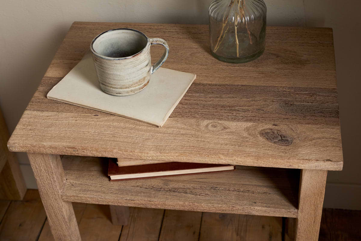The Iya Reclaimed Wood Bedside Table by nkuku holds a ceramic mug on a closed notebook, with another book on its lower shelf and a glass vase with dried stems in the background—perfect for creating a cozy bedroom setting.