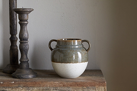 The nkuku Inha Reactive Glaze Vase in Dark Olive & Natural, with two handles and a brown-green glossy top over a speckled off-white base, sits on rustic wood, flanked by two carved dark wooden candle holders against a light gray wall.