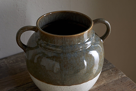 The nkuku Inha Reactive Glaze Vase in Dark Olive & Natural features two handles and sits on a rustic wooden surface, showcasing a glossy dark olive upper half, a matte speckled white base, and smooth texture highlighted by light and shadow.